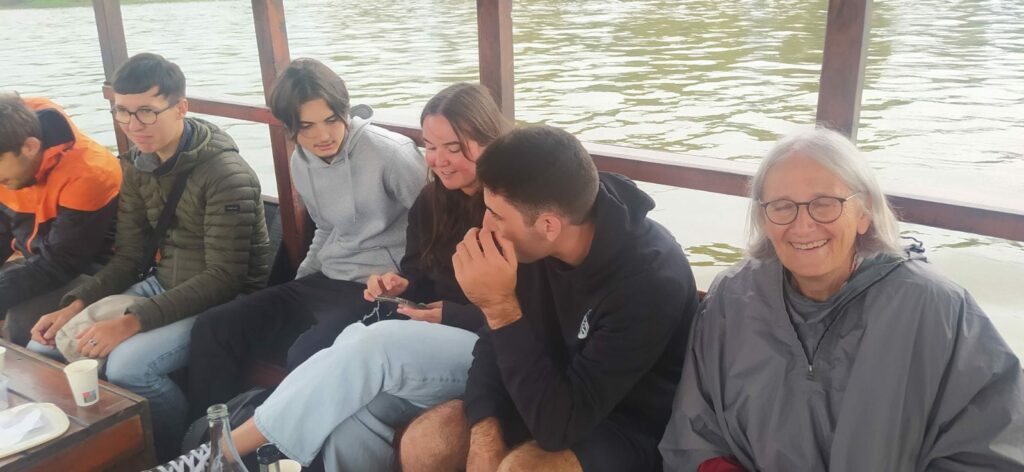 On peut voir 5 jeunes qui discute ensemble assis sur un banc à l'arrière d'un bateau. A leur gauche est assise une personne âgée qui sourit en aillant les yeux fermées 